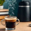 Amazon Plus instant coffee mix review featuring a clear glass cup of black coffee with steam on wooden table